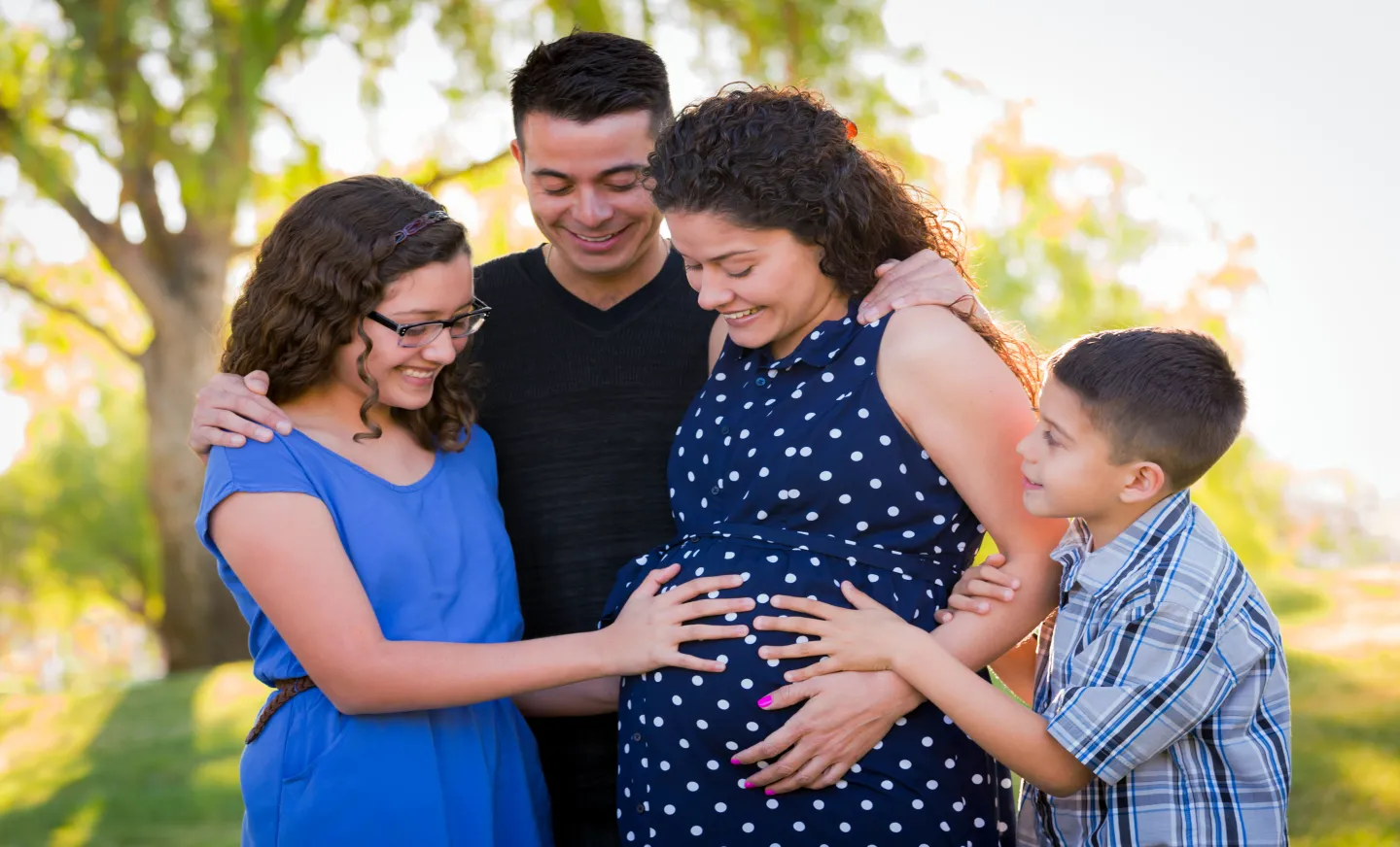 a family, two children and parents are standing outside surrounding the pregnant mother and smiling