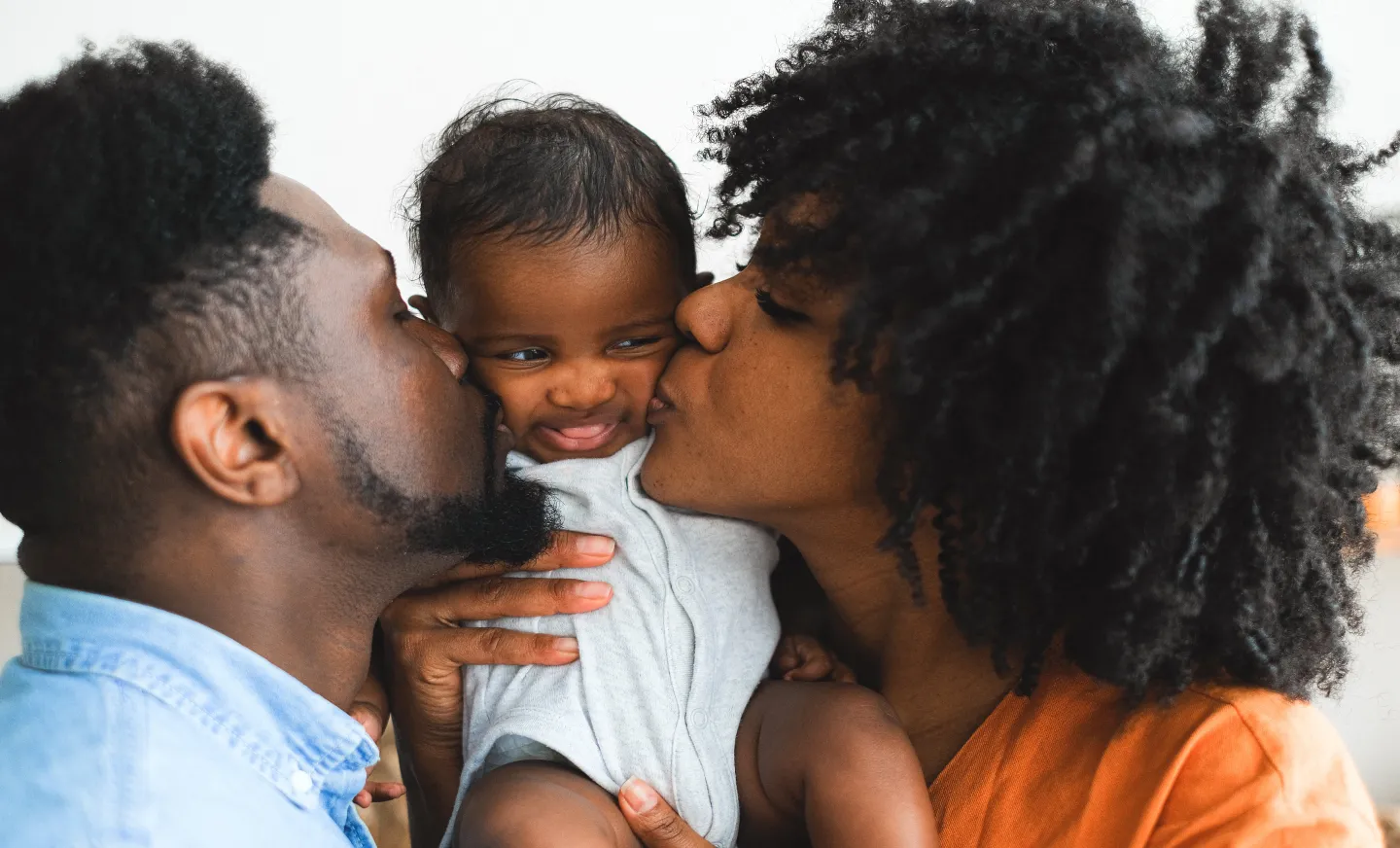 A mom and dad holding their new baby and kissing his cheeks, the baby sticks his tongue out